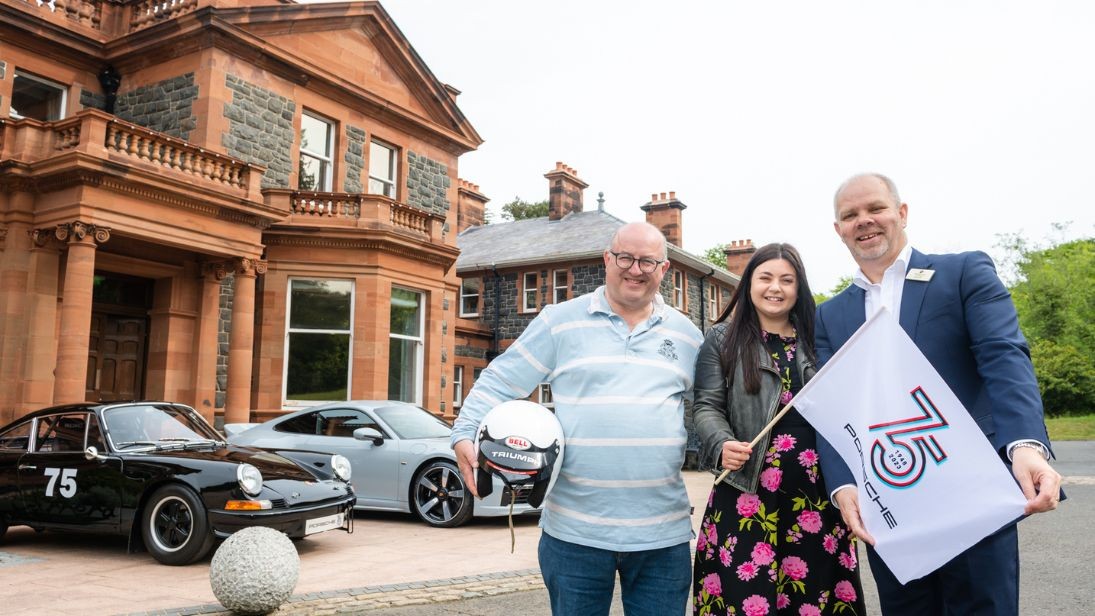 Three people standing in front of a manor house with Porsche cars parked outside. Person on the left is holding a motor racing helmet under his arm, dressed in jeans and a polo shirt. The person in the middle has long dark hair, wearing a floral dress and hold a white flag that says 75 Porsche. The person on the right is wearing a suit and holding the end of the flag.