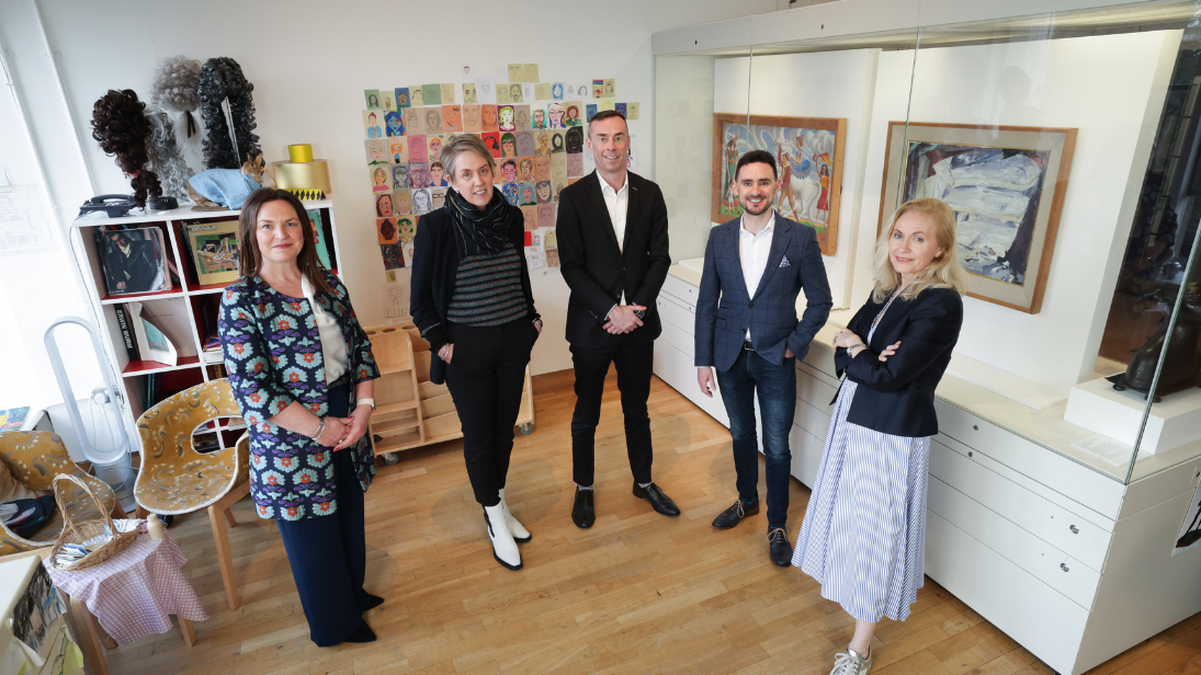 Three women and two men smartly dressed standing in a semi circle inside the Discover Art area of Ulster Museum. The first woman on the left has dark hair and her hands held together in front of her. The second woman has short hair, dressed in all black and has her hands in her pockets. The man in the middle is wearing a suit with his hands held in front of him. The other man next to him has one hand in his pockets. The last person, as we move from left to right, is a woman standing with her arms folded.