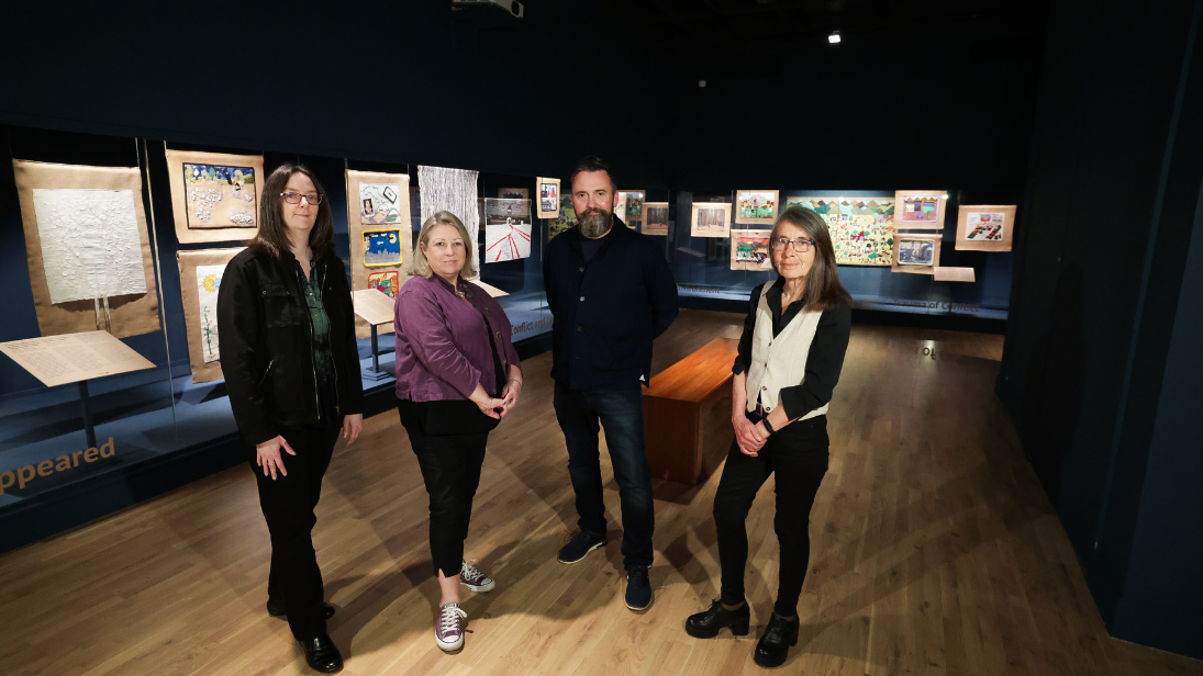 Four people standing inside a dark gallery (which is darkly lit also), with textiles art hanging in glass cases in the background.ay in the Ulster Museum.