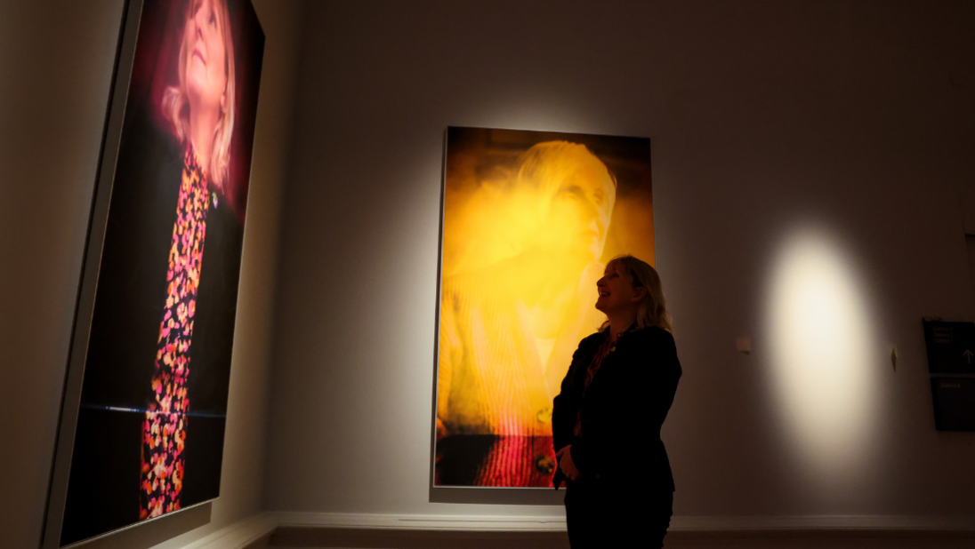 Sandra Peake standing inside the gallery looking up at her photo portrait as part of the 'Principled and Revolutionary: Northern Ireland’s Peace Women' exhibition by Hannah Starkey. There are light and shadow contrasts on the wall.