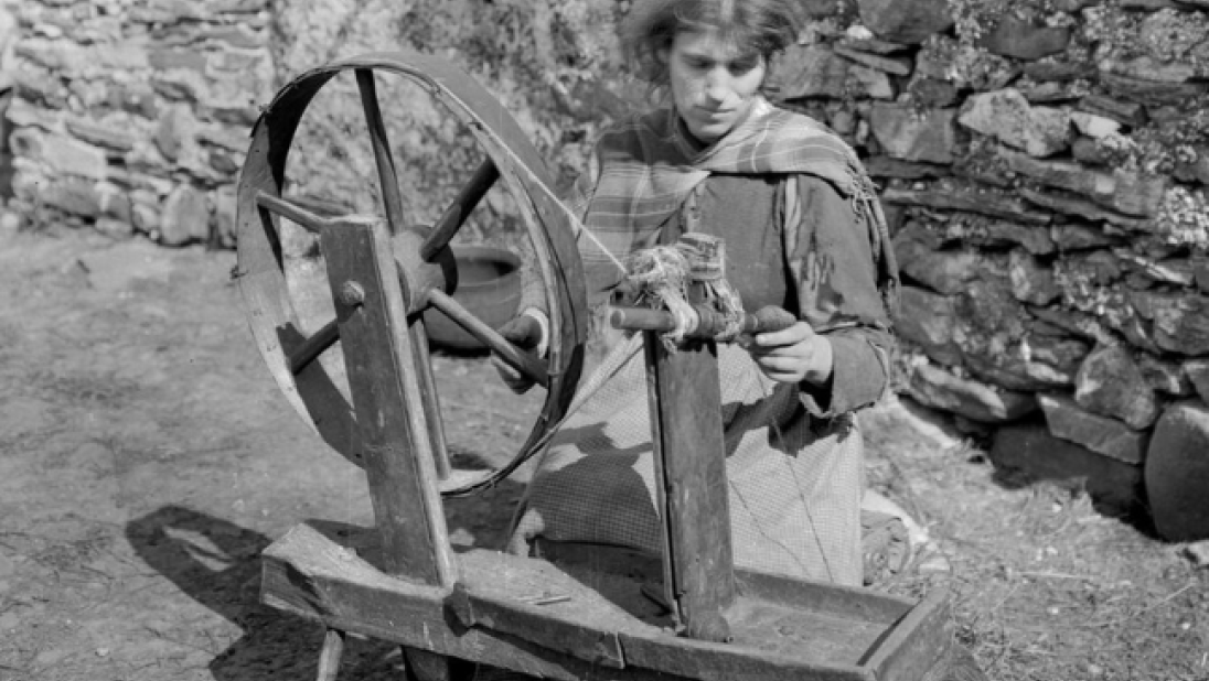 Black and white photograph of a young woman using a spinning wheel outside in rural Ireland