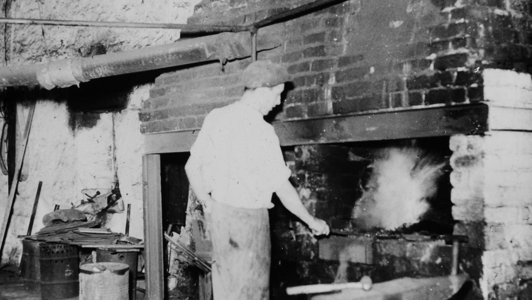 A man stands with his back to the camera. He is by the forge fire, holding a metal object.