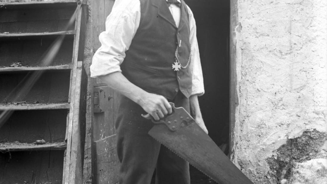 A very tall man stands posing for the camera holding a large saw over a plank of wood.