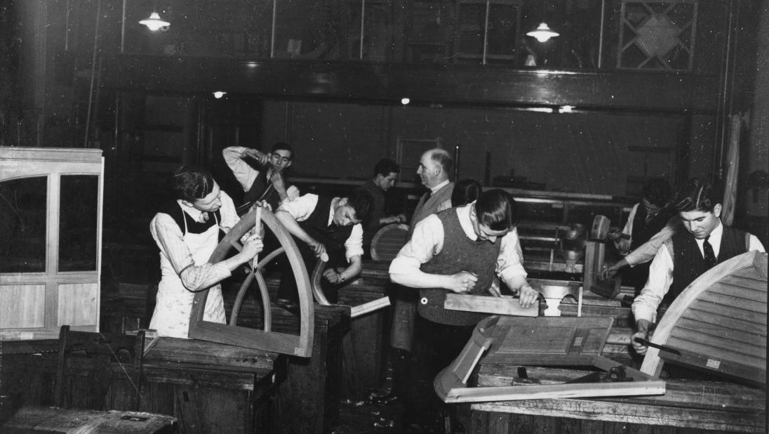 A group of students dressed in suits stand in a woodworking workship, one holding a window frame.