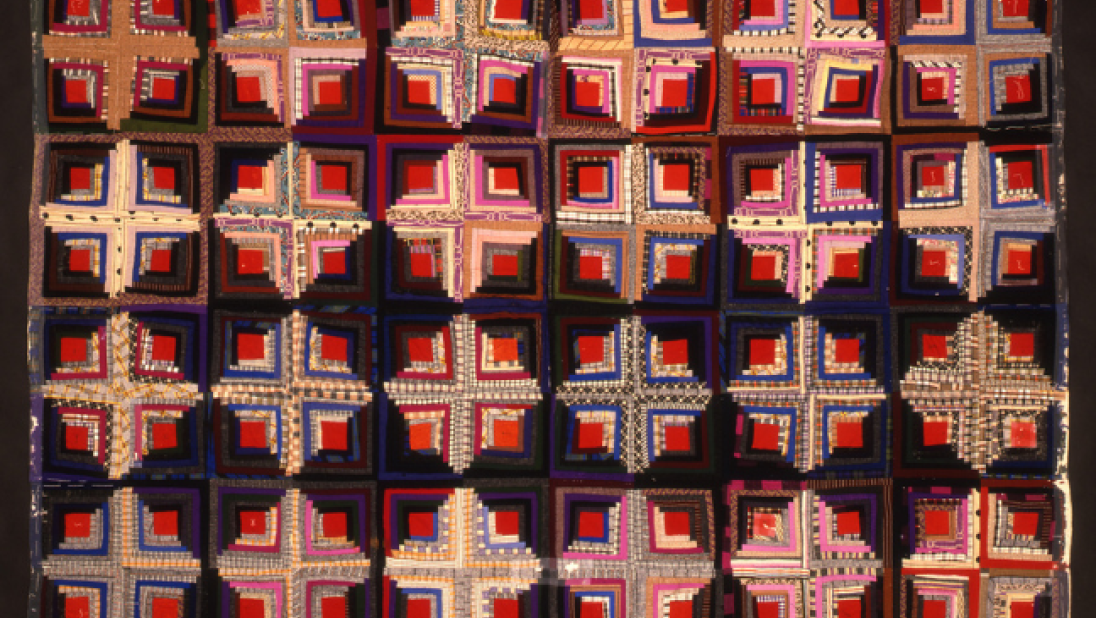 A dark-coloured patterned quilt, with lots of cross, diamond and square patterns.
