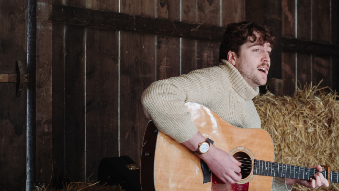 A brunette man in a white jumper playing guitar in a barn
