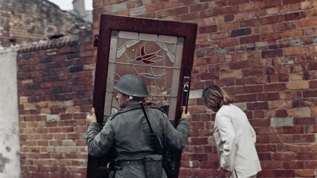 British soldier carrying a door, Bombay Street, West Belfast, Northern Ireland, 1969. A woman in a white jacket and knee-length skirt walks beside him on the footpath.