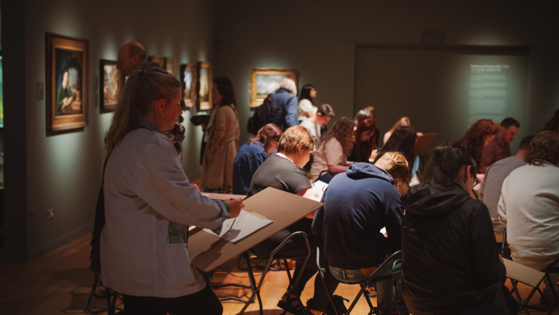 Lots of people in an art gallery taking part in life drawing. One participant is close to camera shot and you can see their page on their board.