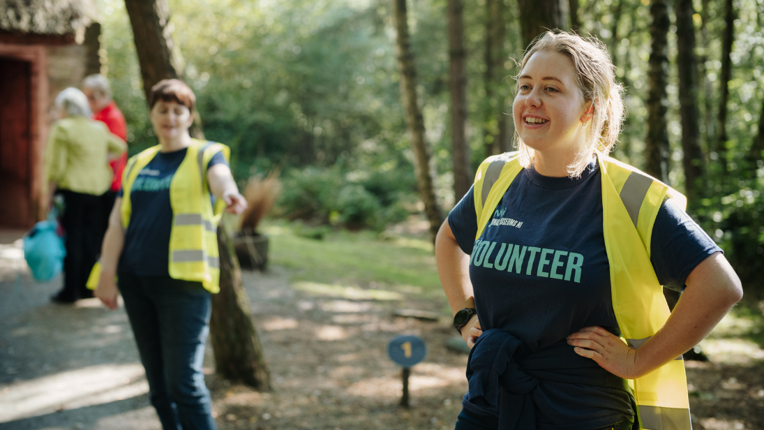 A women in a blue tshirt and yellow high vis jacket standing in a wooded area, smiling