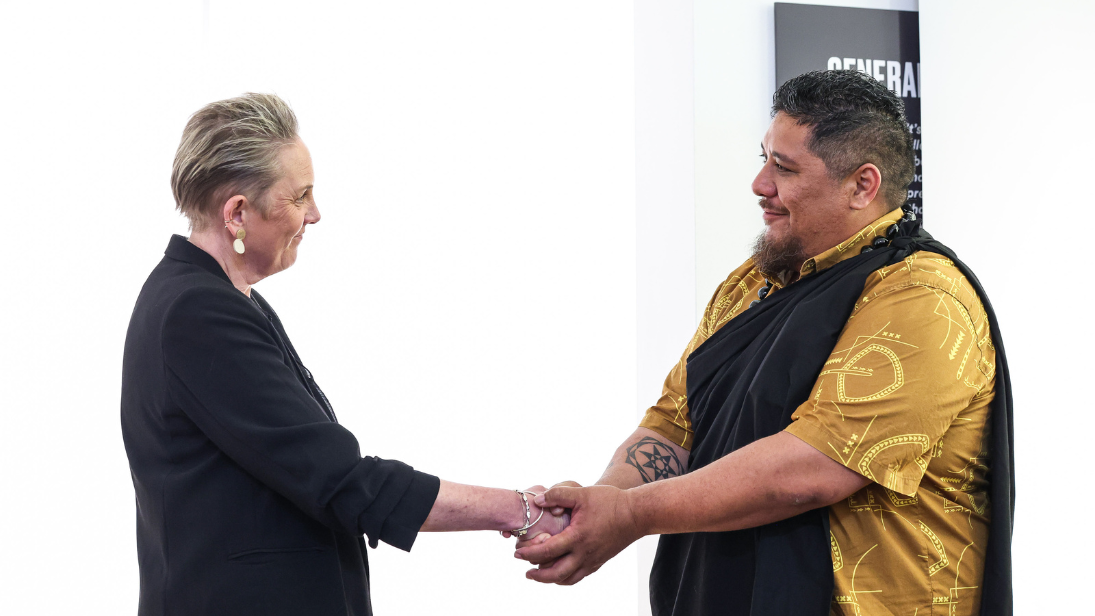 The Chief Executive of National Museums NI dressed in back shakes hands with a delegate from the Office of Hawaiian Affairs and Hui Iwi Kuamoʻo, who is dressed in traditional clothing (black and yellow) in a repatriation ceremony at Ulster Museum.
