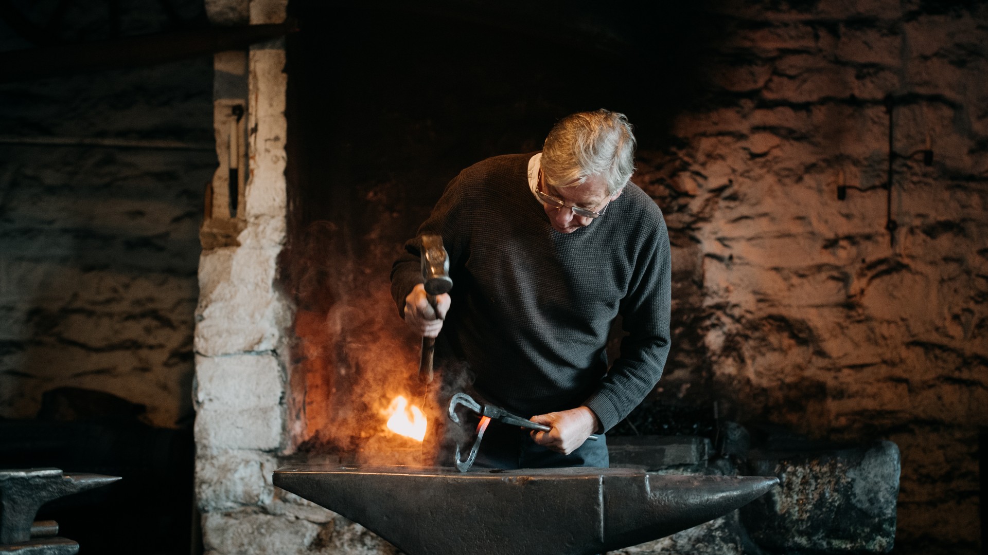 A blacksmith stands behind an anvil with a hammer in his hand. He is about to hammer a piece of metal.
