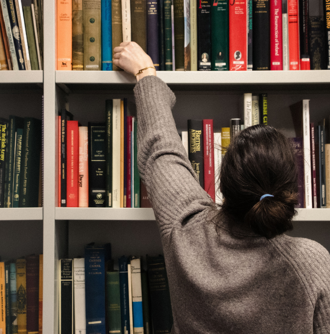 Woman with beige jumper and brown hair, tied up, reaching for a book from a book case