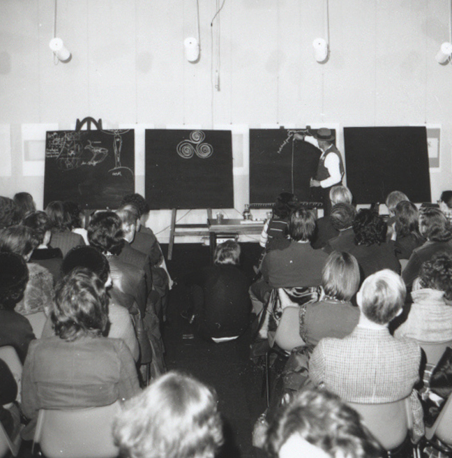 A black and white photograph of a man giving a lecture. He has four blackboards.