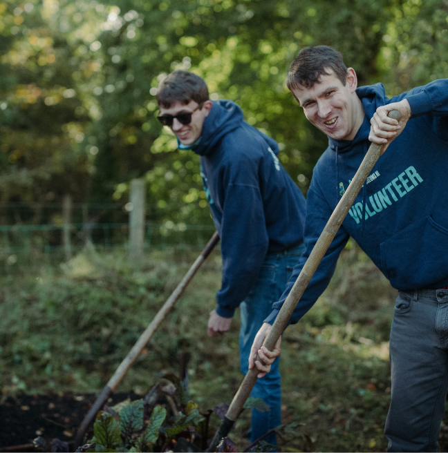Two men stand with gardening tools in a lush green garden.