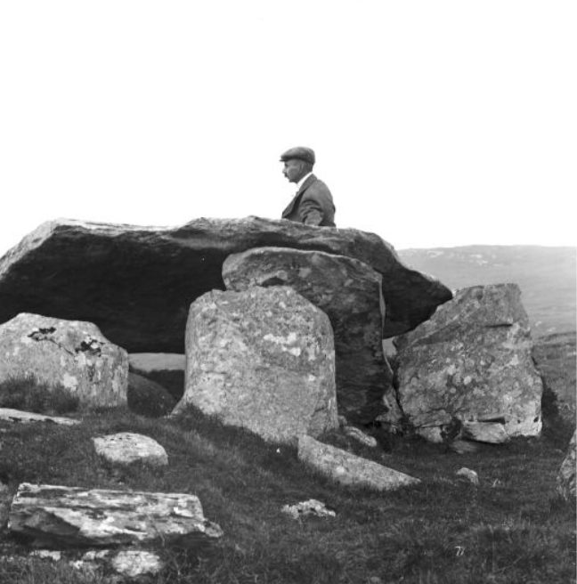 A black and white image of a man standing beside an archeological stone feature.