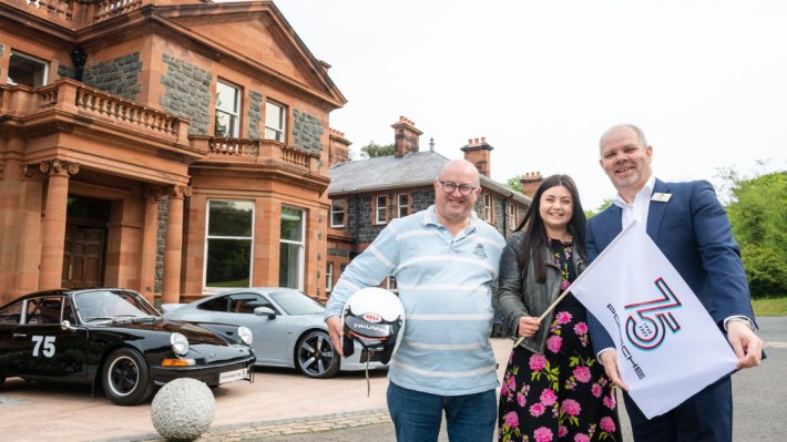Three people standing in front of a manor house with Porsche cars parked outside. Person on the left is holding a motor racing helmet under his arm, dressed in jeans and a polo shirt. The person in the middle has long dark hair, wearing a floral dress and hold a white flag that says 75 Porsche. The person on the right is wearing a suit and holding the end of the flag.