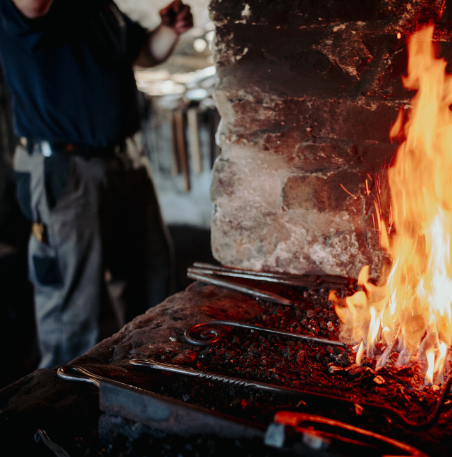 A blacksmith in his forge in the backgroun; a fire and tools in the foreground.
