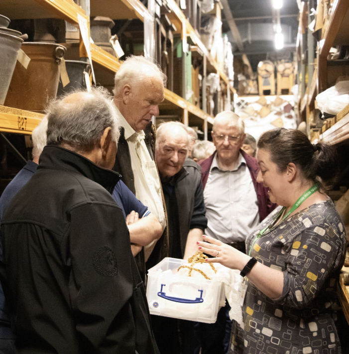 A group of men stand around a curator, who is holding a box and speaking about the woven object inside.