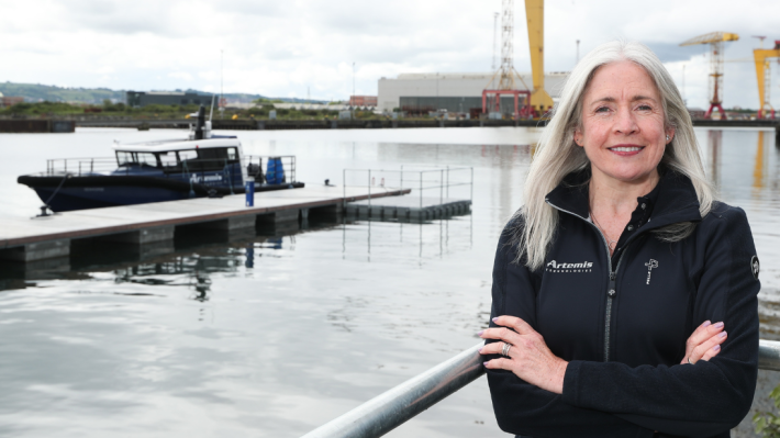 Professor Katrina Thompson standing with her arms crossed in Belfast harbour, Harland and Wolff cranes visible in the background