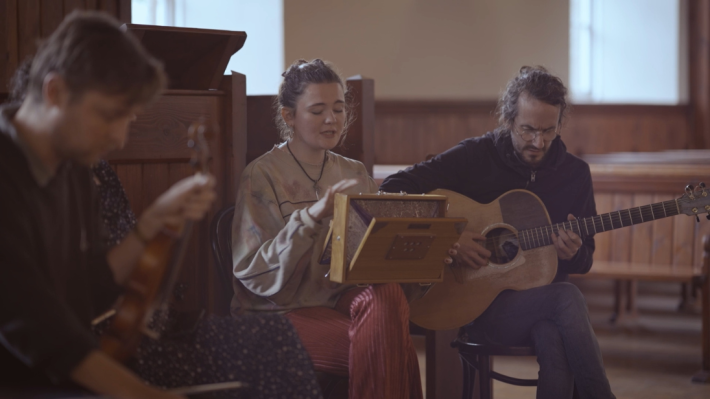 A group of musicians sit in a semi circle in a church, playing their instruments. 