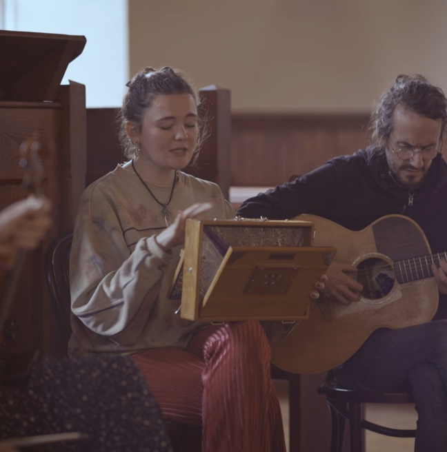 Musicians sit in a semi-circle with their instruments, including a guitar and a fiddle just out of frame. They are in an old church.