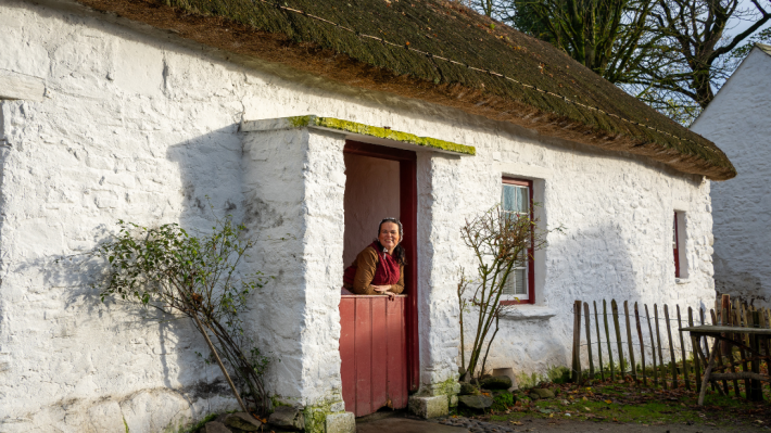 A woman stands in the doorway of a thatched cottage. She looks out at us, smiling.