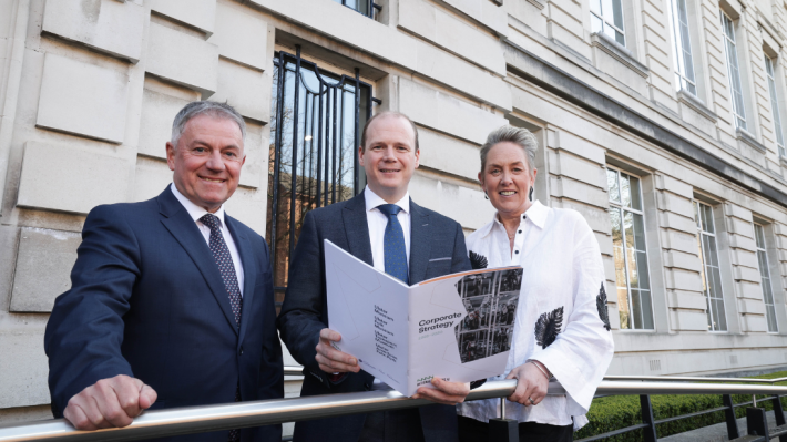 Three people standing beside a railing outside the Ulster Museum, the white-grey stone of the building visible in the background, with the person in the middle holding an A4 document titled Corporate Strategy 2025-2030.