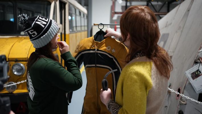 Two women regard a yellow leather motorcycle suit.