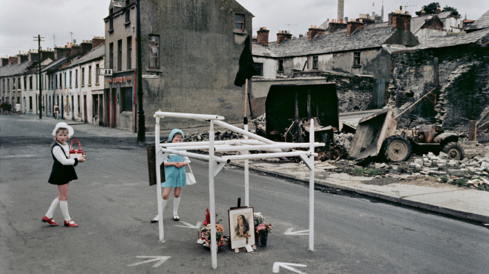 Scene of damaged houses on a street, with two young girls in dresses and hats in the middle of the street beside a 'street memorial' made during the Troubles. ©Estate of Akihiko Okamura / ©佐藤純子