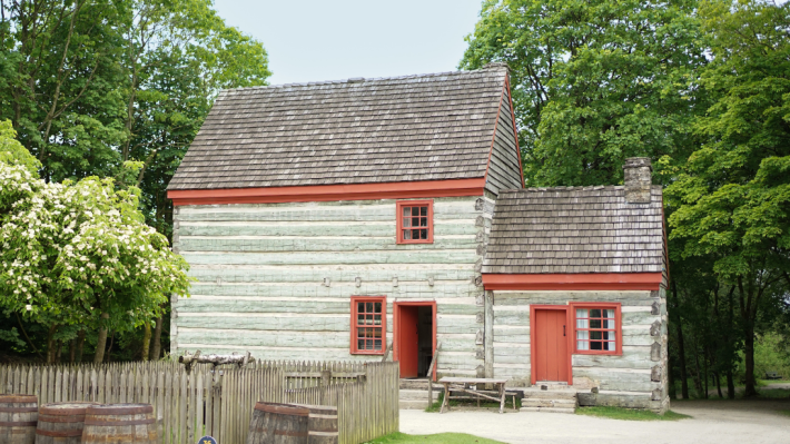 A log farmhouse sits amongst trees. The door and roof trimmings are red, while the logs are a blueish gray.