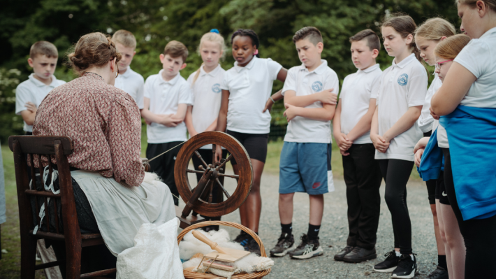 School kids all standing in a casual line, watching a museum demonstrator weaving wool. Their school uniforms are white polo shirts with a blue badge.