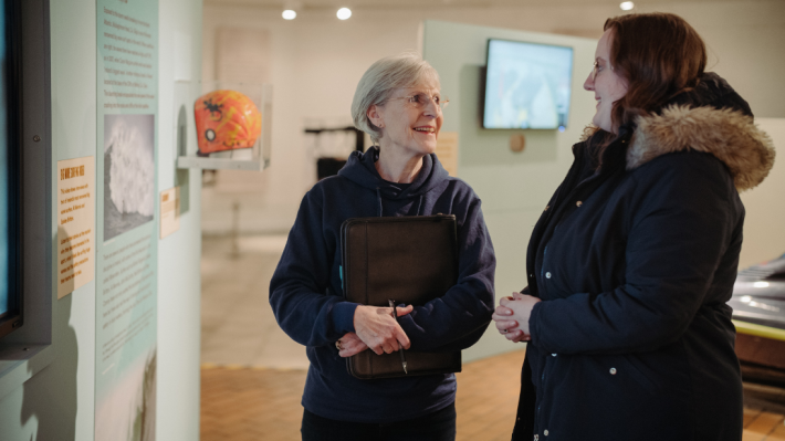 An older lady with glasses, holding a clipboard against her chest, smiling and talking to another lady wearing a cost with a fur trimmed hood inside an exhibition on Irish surfing at the Ulster Transport Museum