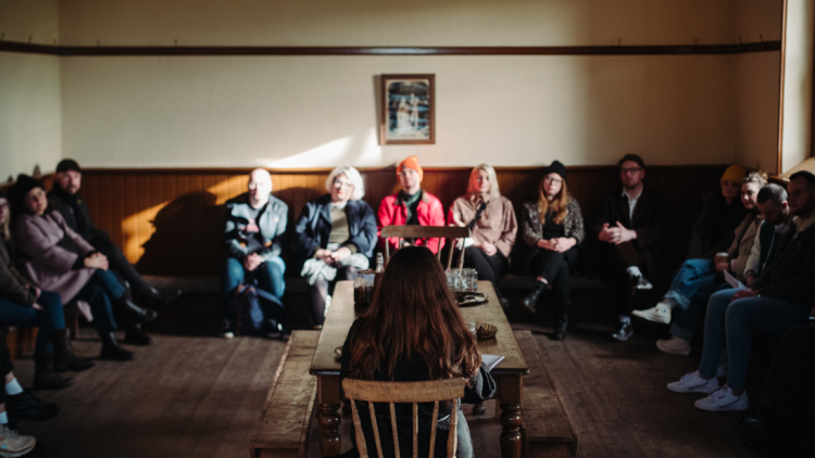 Group of people sitting on benches around a room listening to a poet