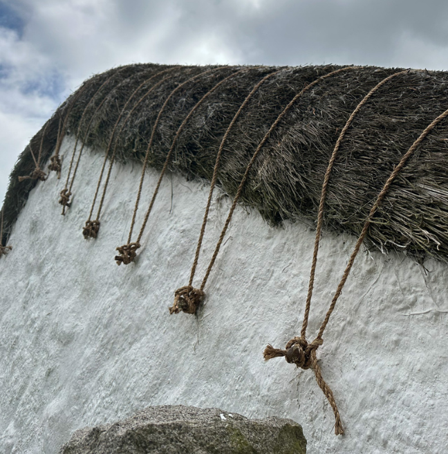 The gable end of a cottage showing ropes tied to metal pegs. The rope holds down thatch on the roof. 