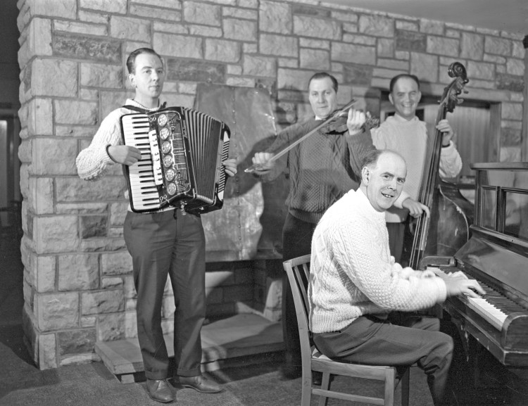 Four musicians pose for a photo. They are playing an accordian, a piano, a fiddle, and what appears to be an upright bass.