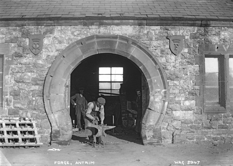 The entrance to a forge, with a man inside staring out. Another man in the doorway is working on an anvil.