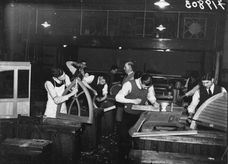 A group of students dressed in suits stand in a woodworking workship, one holding a window frame.