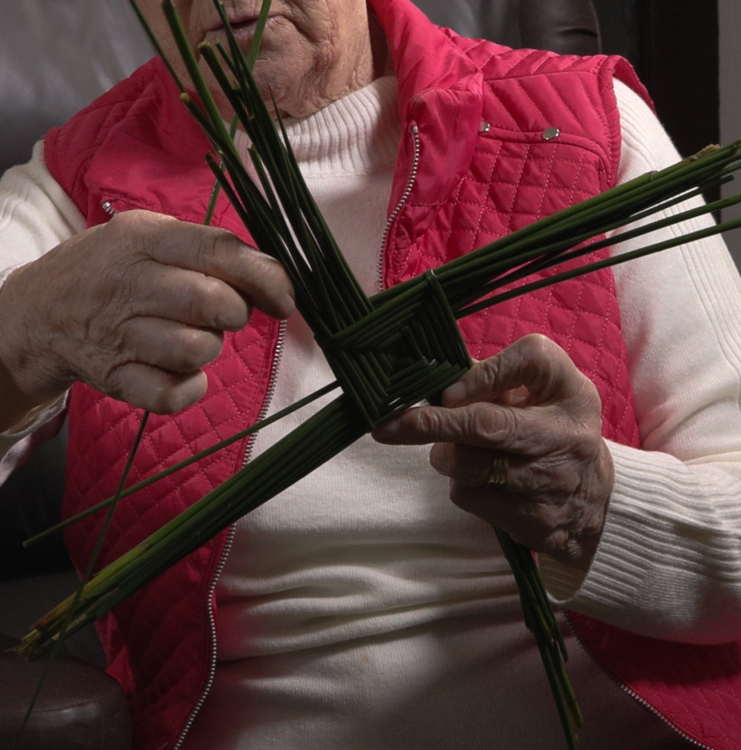 A woman in a red cardigan weaves a St. Brigid's Cross.