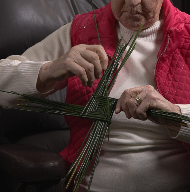 A woman in a red cardigan weaves a St. Brigid's Cross.