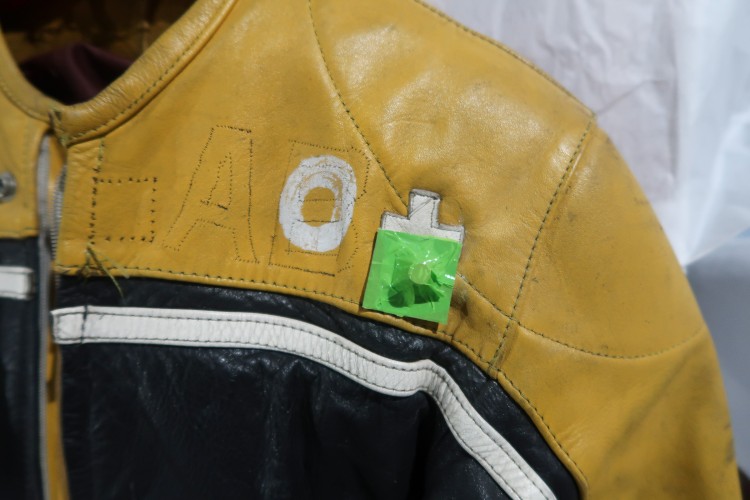A close up of the top of a racing jumpsuit. There are white patches spelling 'O +' sewn on. There is a small magnet on top of the patch.