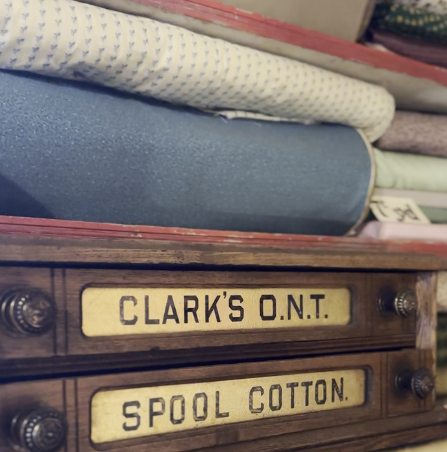 Cotton sheets sit atop one another on a shelf, above a sign reading 'Clark's ONT spool cotton'.