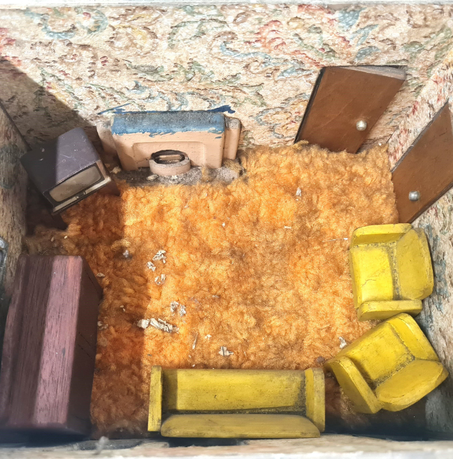 The inside of a dolls' house. The floor is a shaggy orange carpet. It is a living room, with two armchairs, a couch, and an old television in the corner.