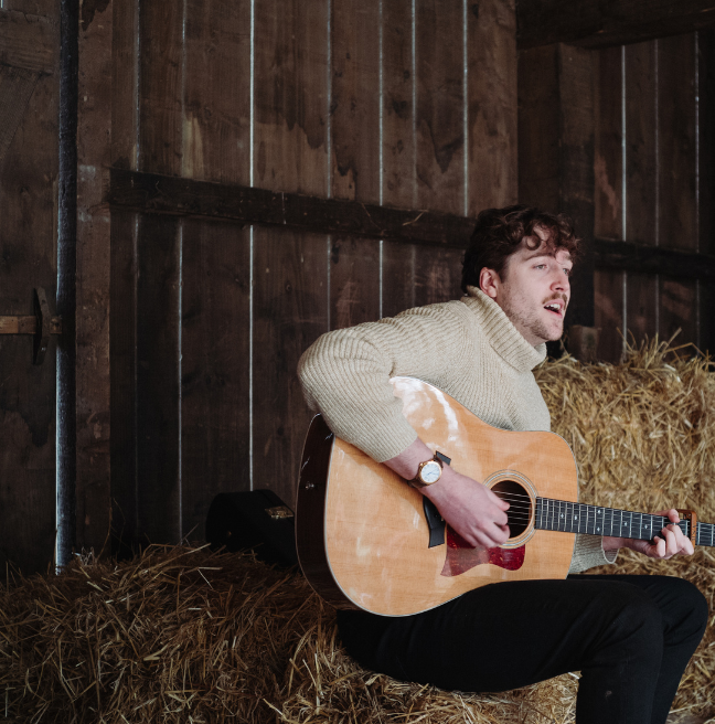 A brunette man in a white jumper playing guitar in a barn