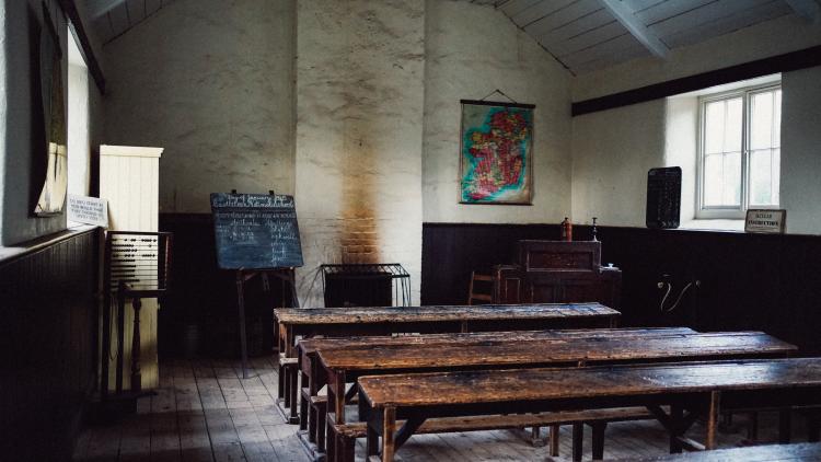 A view of an old schoolhouse. Wooden desks sit in rows facing the teacher's desk and a slate board.