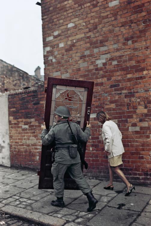 British soldier carrying a door, Bombay Street, West Belfast, Northern Ireland, 1969. A woman in a white jacket and knee-length skirt walks beside him on the footpath.