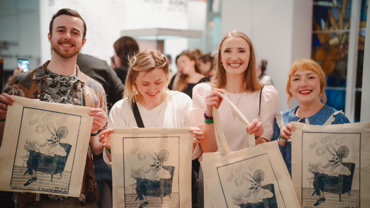Four people standing in a line holding tote bags with a navy ink print on them.