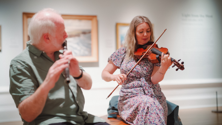 Two Irish Trad musicians playing in an art gallery, the person on the left is playing a whistle, the person on the right plays the violin.