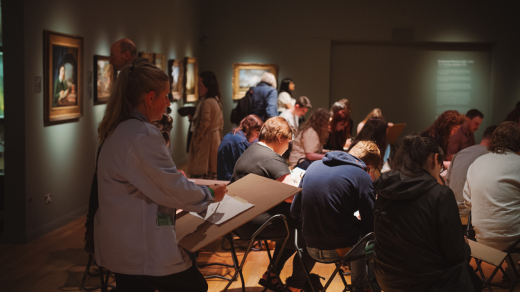 Lots of people in an art gallery taking part in life drawing. One participant is close to camera shot and you can see their page on their board.