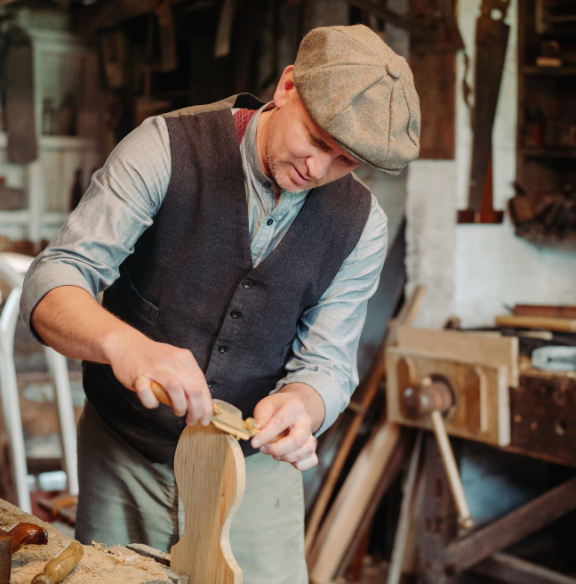 A heritage carpenter in 1900s dress - peaked cap, wool waistcoat, rolled up sleeves shirt - performing wood working skills.