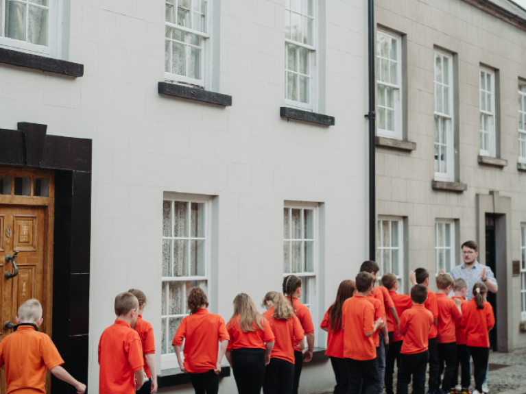 Primary school pupils wearing red tops and black bottoms lined up in single file beside houses at the Ulster Folk Museum with the education facilitator (wearing glasses and wearing a white top) at the top of the line